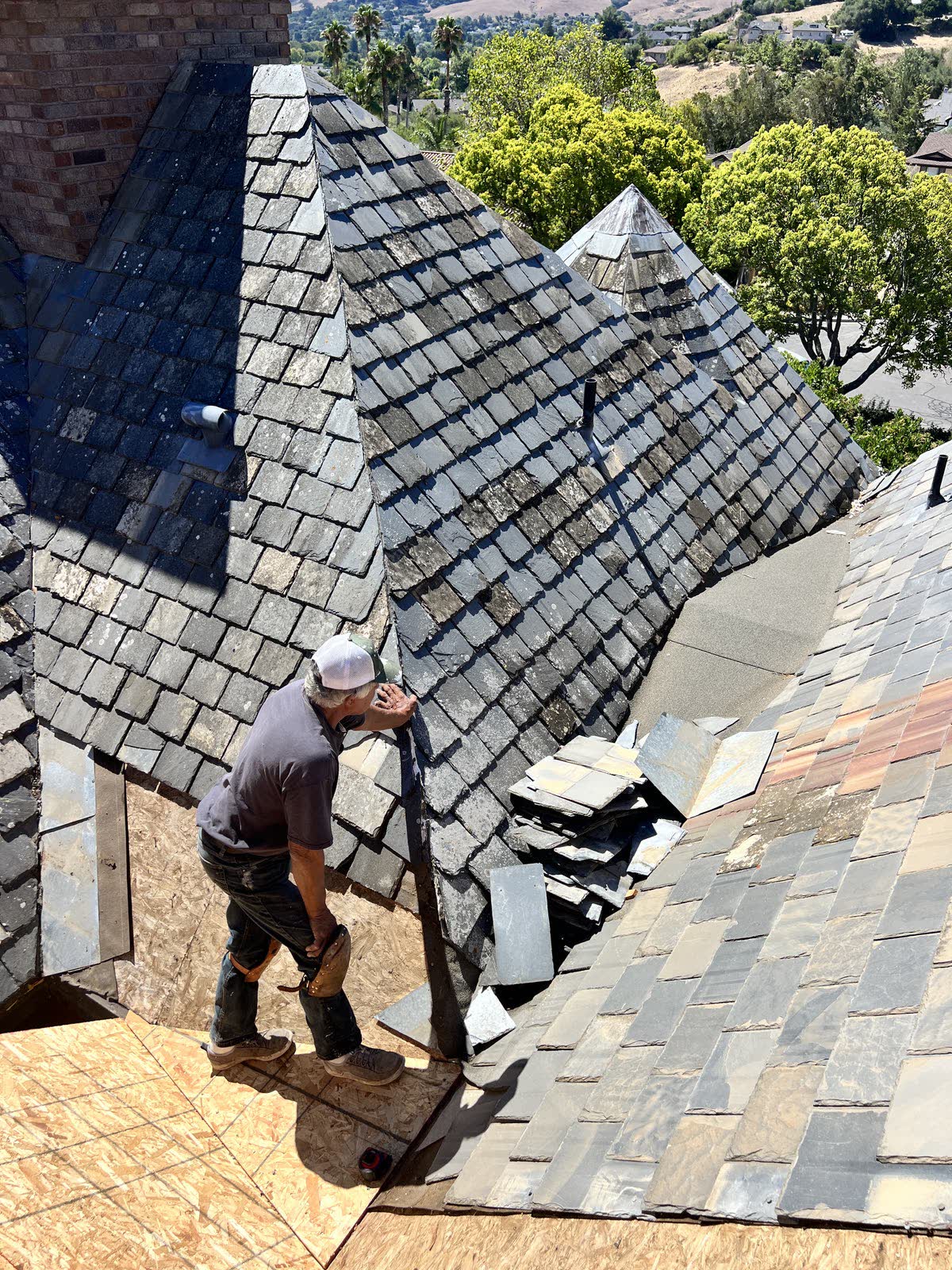 Looking down on a multi-hip roof being covered in natural slate. A man in a
                grey shirt, white cap and tool belt stands on freshly laid sheathing at a
                valley, setting slates by hand up the hip. Loose slates are stacked on the
                deck beside him, a brick chimney rises at the left, and oaks and dry hills
                run away behind the house.