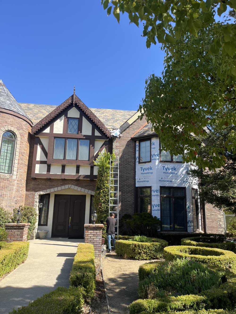 The front of a large Tudor Revival house from the driveway: brick and
                  half-timbering, a steep slated gable with a finial, a round turret at the
                  left, clipped boxwood hedging and brick entry piers. A ladder stands
                  against the wall with a man at its foot.