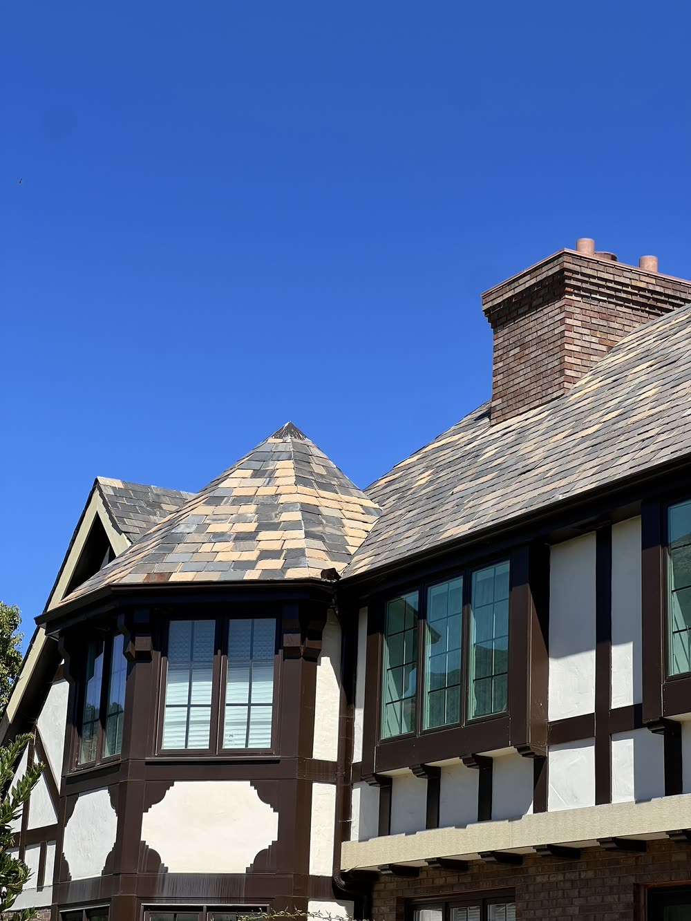 A conical turret roof over a bay window on a Tudor Revival house, covered
                  in graduated slate that runs from grey through tan to purple, against a
                  deep blue sky. A brick chimney stands behind it and the timber-framed
                  gable and leaded windows sit below.